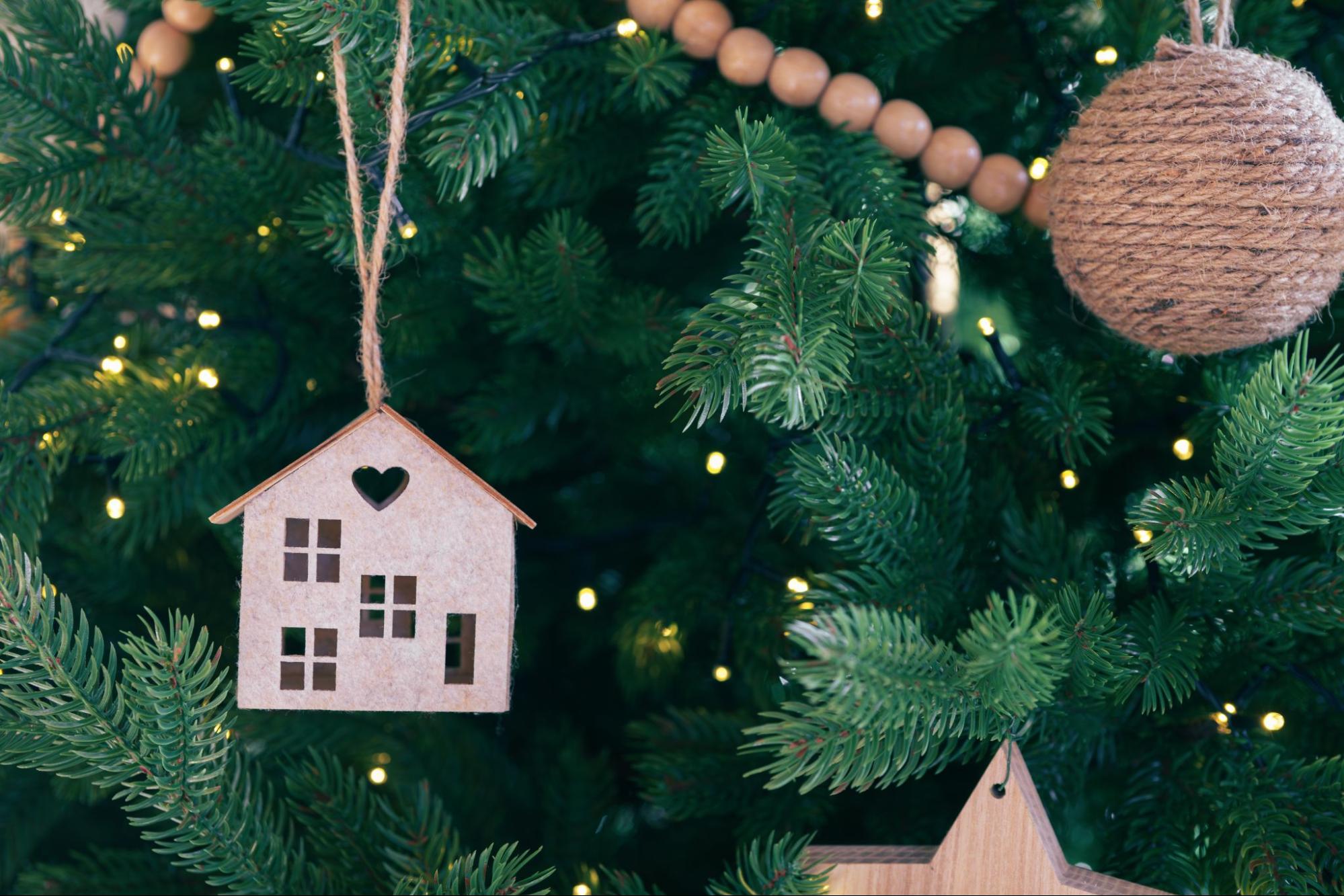 Handcrafted wooden house ornament with heart cutout hanging on Christmas tree branch with warm holiday lights in the background. ©Katerina Maksymenko