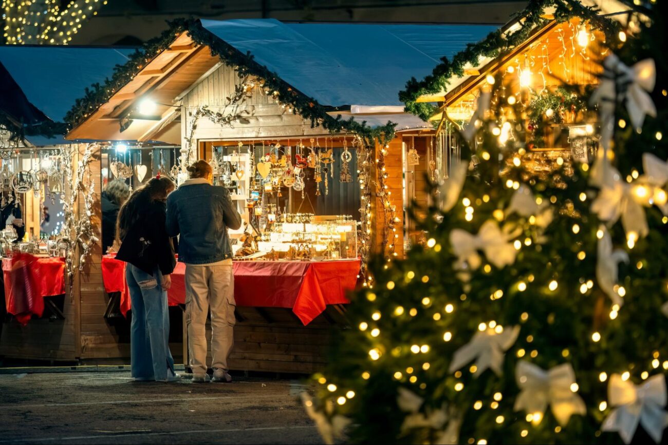 Couple exploring festive wooden market stalls at a Christmas tree-lit market in DFW ©Rostislav Glinsky
