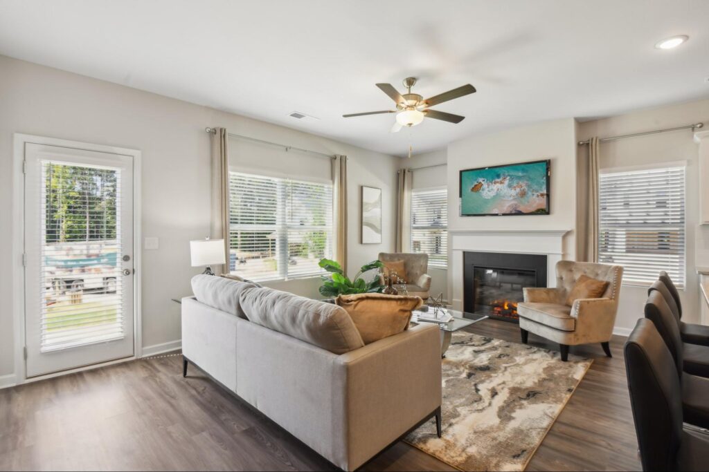 A bright, open-concept living room featuring a modern fireplace, neutral-toned furniture, and large windows that let in natural light. The space is styled with a plush rug, a ceiling fan, and views leading toward the dining area.