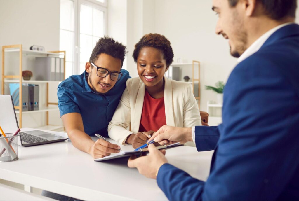 A happy young couple sits at a table, smiling as they sign homeownership documents. A professional real estate agent sits across from them, guiding them through the final steps of the closing process. ©Studio Romantic